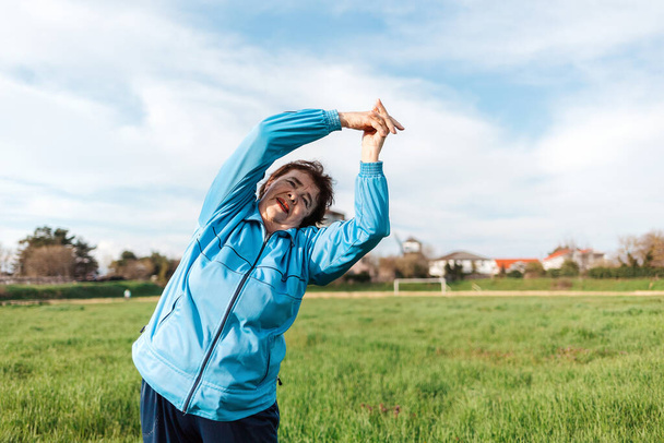 Wellness and sports. Smiling elderly woman in sports clothes, doing exercise in the park. Tilts to the side. International Day of Older Persons. - Foto, afbeelding