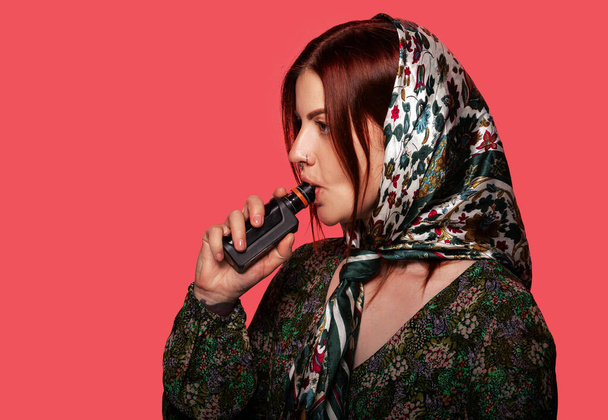 Γυναίκα καπνίζει ηλεκτρονικό τσιγάρο με τρυπημένη μύτη και μοδάτη μαντίλα. Κόκκινο φόντο, χώρος για κείμενο. Ομορφιά, μόδα έννοια. - Φωτογραφία, εικόνα
