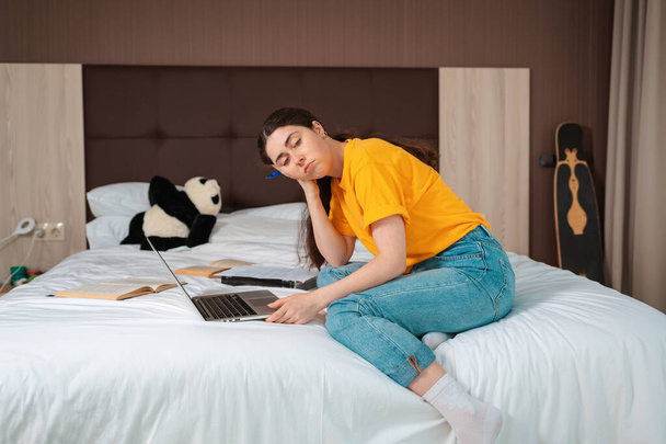 Concept of online courses. A young Caucasian woman is sitting with a bored look on the bed, and a laptop and books are lying next to her. Home interior. - 写真・画像