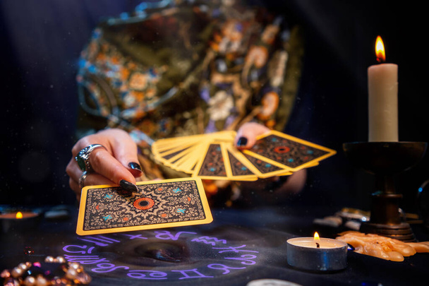 A cartomante segura um fã de cartas em suas mãos e segura um cartão com uma mão. O círculo astrológico brilha à mão. Close-up. O conceito de adivinhação, magia e esoterismo. - Foto, Imagem
