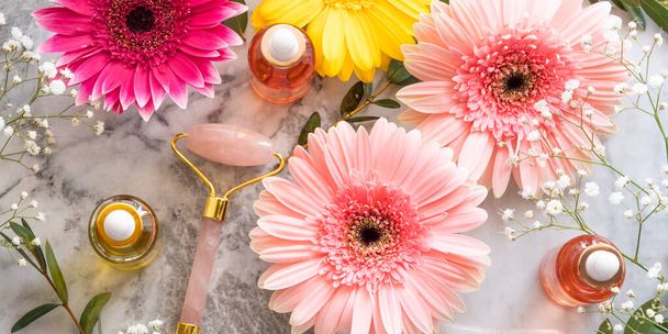 Rózsaszín kvarc arc görgő gua sha és szérum palackban márvány asztalon. Bőrápolási koncepció virágokkal - Fotó, kép