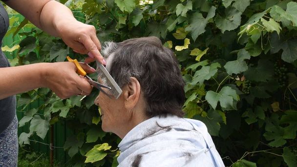 Hände schneiden Haare an der Schläfe einer alten Dame mit halbgrauem Kurzhaarschnitt - Foto, Bild