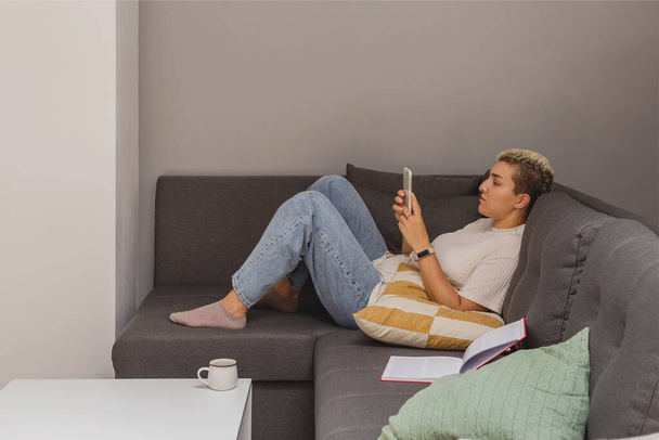 Egy nő fekszik egy kanapén egy okostelefonnal egy minimalista otthon belsejében. - Fotó, kép