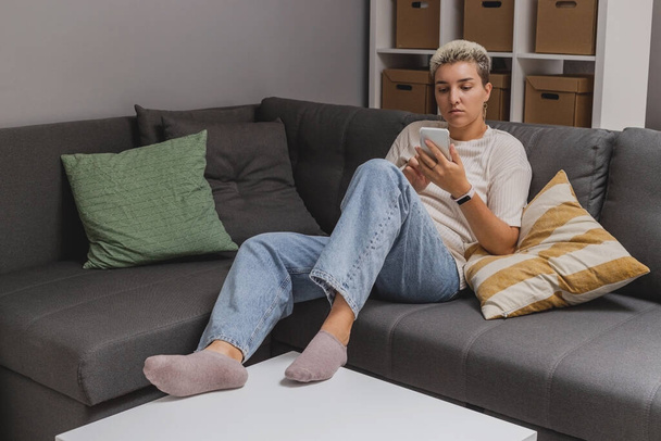 A woman lies on a sofa with a smartphone in a minimalistic home interior. - Φωτογραφία, εικόνα
