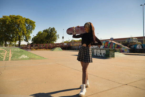 Молодая красивая девушка с панк-стилем держа скейтборд через плечо и с одной рукой на талии глядя на бесконечность в скейтборд-парке. - Фото, изображение