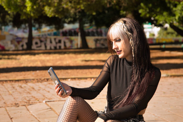 Punk tarzı genç ve güzel bir kız yerde oturmuş cep telefonundan sosyal ağları kontrol ediyor.. - Fotoğraf, Görsel