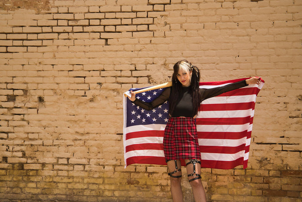 Pretty young girl with pigtails and punk style with a baseball bat behind her neck and a U.S. flag behind her back with a yellow wall in the background. - Photo, Image