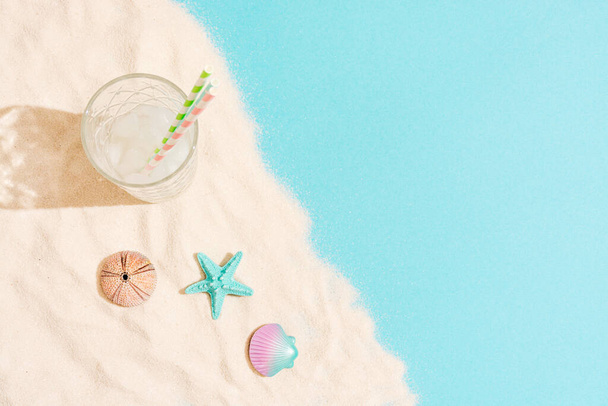 Summer concept composed of tall glass with ice and straws and of sea urchin, starfish and a shell laid out on sand and blue background. - Photo, Image