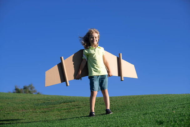 Child playing with cardboard toy wings in the park. Concept of children day. Kid boy in an pilot costume is playing and dreaming of becoming a aviator spaceman. - Foto, afbeelding