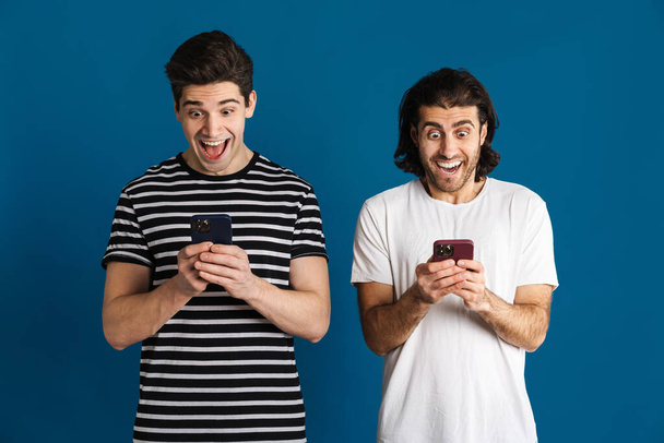 Excited two men screaming while using cellphones isolated over blue background - Photo, image