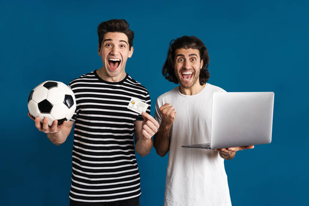Excited two men using laptop while holding football and credit card isolated over blue background - Foto, imagen