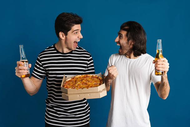 White excited two men screaming while drinking beer and eating pizza isolated over blue background - Fotografie, Obrázek