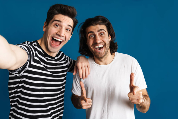 White excited two men pointing finger at camera while taking selfie photo isolated over blue background - Fotografie, Obrázek