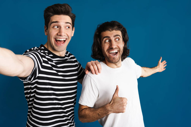 White excited two men gesturing thumb up while taking selfie photo isolated over blue background - Фото, изображение