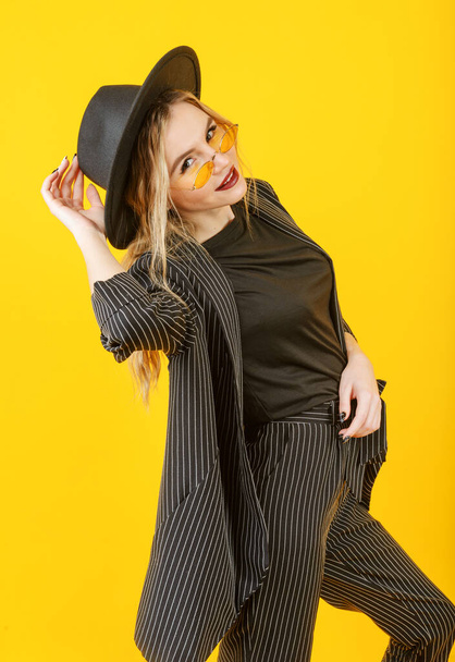 young blonde woman in a black jacket ,glasses and hat smiles on a yellow background  - Фото, зображення