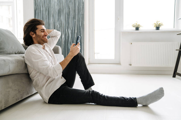 Smiling mid aged turkish man holding mobile phone sitting on a floor in the living room - 写真・画像