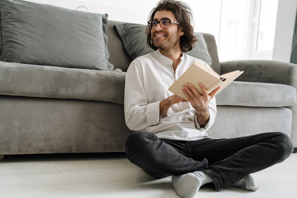Smiling mid aged brunette turkish man sitting on a floor reading book in the living room - Φωτογραφία, εικόνα