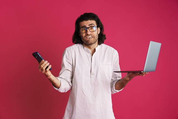 Unshaven confused man posing with mobile phone and laptop isolated over pink background - Photo, image