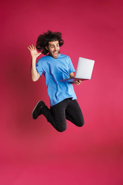 Excited brunette man gesturing and using laptop while jumping isolated over pink background - 写真・画像