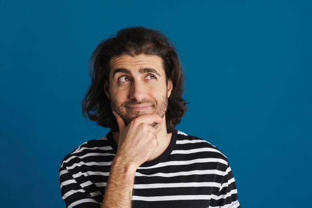 Unshaven brunette man in striped t-shirt thinking and looking aside isolated over blue background - Foto, immagini