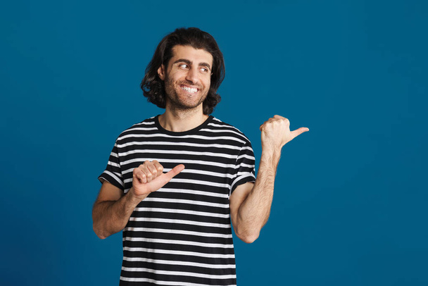 Unshaven brunette man smiling and pointing fingers aside isolated over blue background - 写真・画像