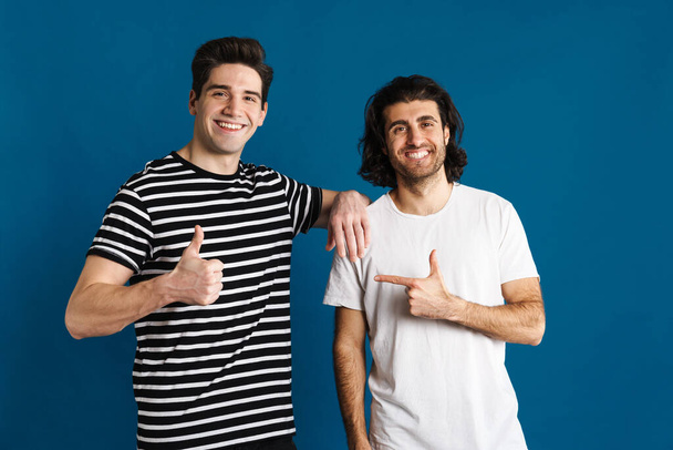 White smiling two men showing thumb up and pointing finger aside isolated over blue background - Foto, Imagem