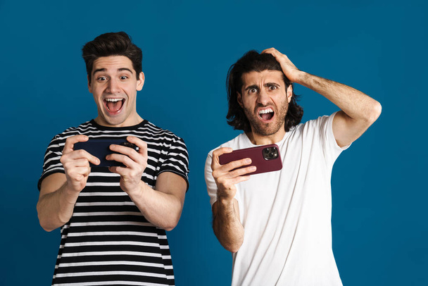White brunette two men playing online game on cellphone isolated over blue background - Fotó, kép