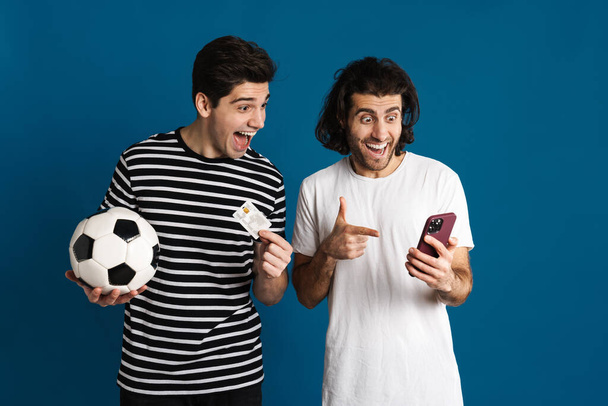 Excited two men using cellphone while holding football and credit card isolated over blue background - Foto, immagini