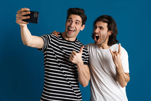 White excited two men taking selfie on cellphone while gesturing isolated over blue background - Foto, immagini