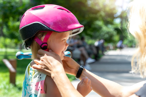 Close-up mom parent hand put on and fasten safety helmet on cute blond caucasian toddler girl for riding bike or scooter city street park outdoors on summer day. Child sport activity protection care - Photo, Image