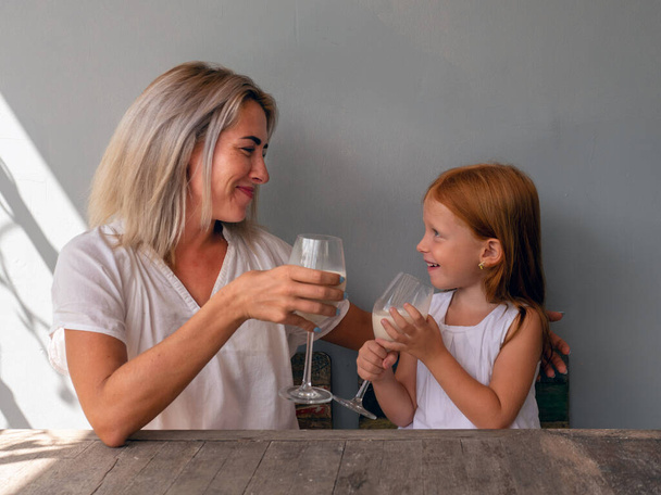 Mutter und sechsjährige rothaarige Tochter trinken am Tisch Milch aus Gläsern - Foto, Bild