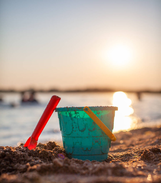 Kinderspeelgoed om op het zand te spelen. Plastic emmer en hark op het strand bij zonsondergang. Het concept van zomer, gezinsvakantie en vakantie. - Foto, afbeelding