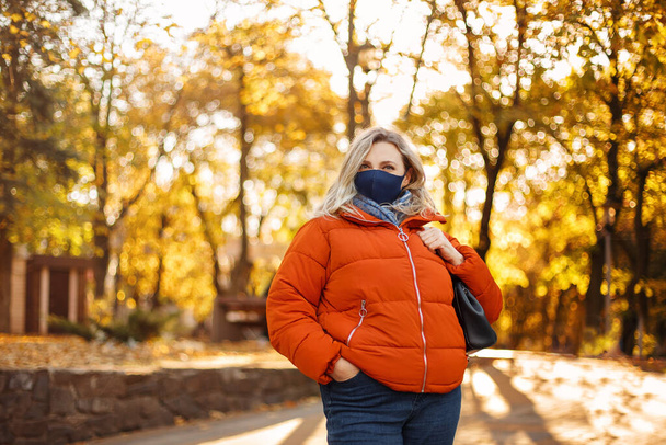 Sonbahar parkında güneşli bir günde salgın sırasında asfalt yolda yürüyen maske takan sarışın kadın. - Fotoğraf, Görsel