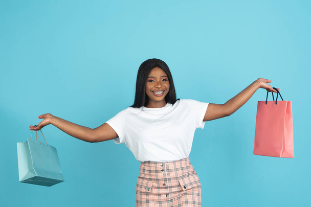 Portrait of smiling African young woman with paper bags isolated on blue studio background. Concept of human emotions, facial expression, youth, sales, ad. - Fotoğraf, Görsel
