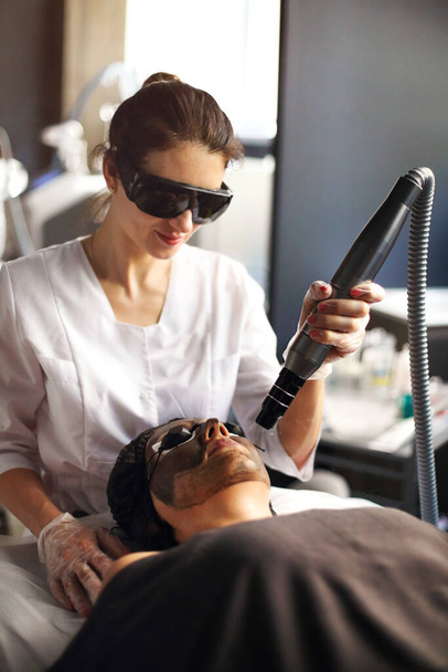 Kosmetyczka trzymająca laser piękności w pobliżu twarzy kobiety pokrytej maską węglową i okularami ochronnymi, zabiegi spa z laserowymi i czarnymi maskami - Zdjęcie, obraz