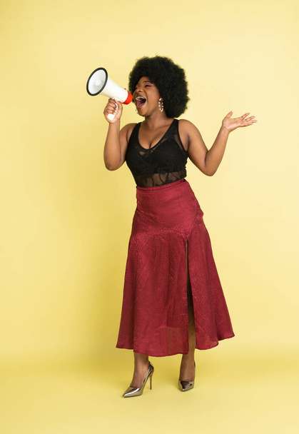 Portrait of African young woman shouting at megaphone isolated over light yellow studio background. Concept of human emotions, facial expression, youth, sales, ad. - Фото, зображення