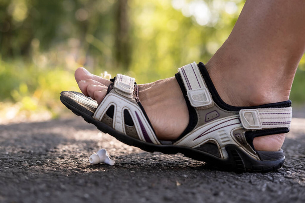 Nainen jalka sandaalit astuu nuhjuinen langattomat kuulokkeet, joka sijaitsee asfaltti jalkakäytävä, ulkona, vastaan hämärtynyt tausta. - Valokuva, kuva