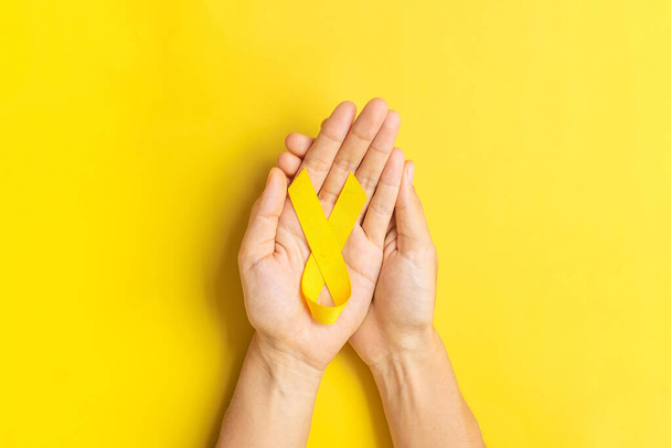 kädestä tilalla keltainen nauha keltaisella pohjalla elämisen ja sairauden tukemiseksi. Syyskuu Itsemurhien ehkäisypäivä, Lapsuuden syöpätietoisuuskuukausi ja Maailman syöpäpäivän käsite - Valokuva, kuva