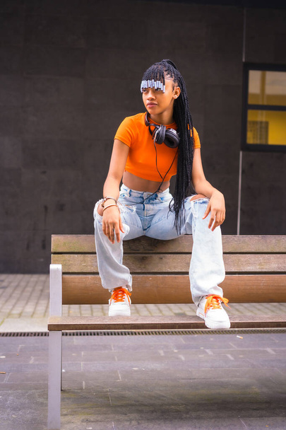 Lifestyle with a young trap dancer in the city. Black grind girl of African ethnic group with orange t-shirt and cowboy pants, with headphones on a black background - Foto, Imagem