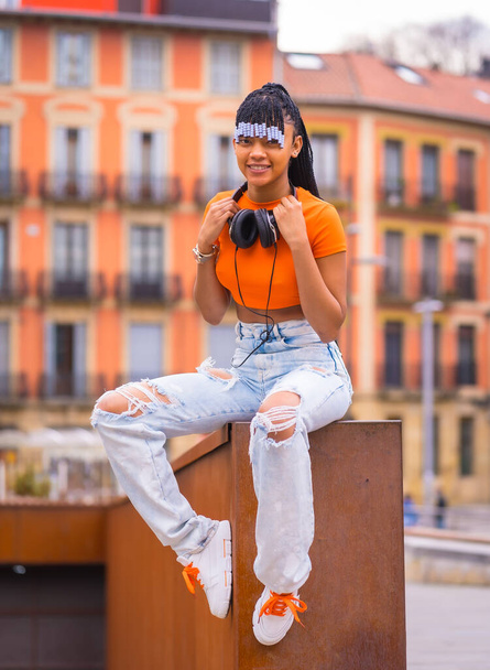 Lifestyle with a young trap dancer with braids. Black-race girl of African ethnic group with orange shirt and cowboy pants sitting. With orange houses background - Φωτογραφία, εικόνα