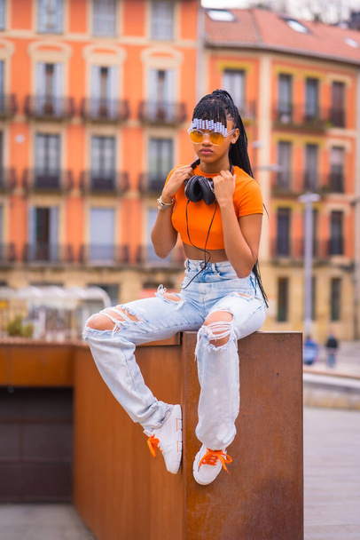 A young trap dancer with braids. She pose from the black grinding girl of African ethnic group with orange shirt and cowboy pants sitting. With orange houses background - Foto, immagini