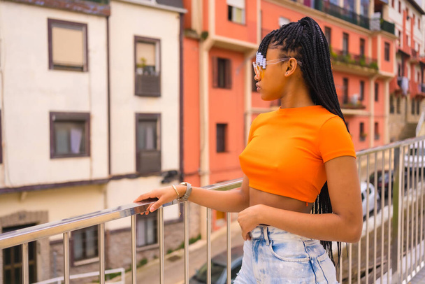 A young trap dancer with braids. Black ethnic girl with orange t-shirt and cowboy pants with a cup Take-Away in the city - 写真・画像