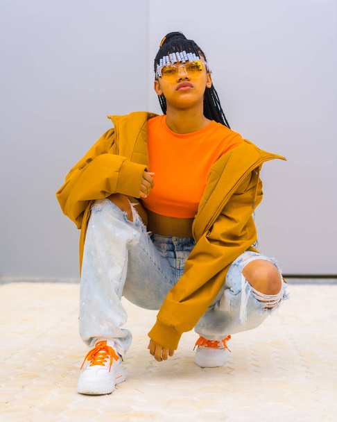A young trap dancer with braids. Black ethnic girl with t-shirt, orange sunglasses and a jacket, crouched looking at camera with seductive face - Photo, Image