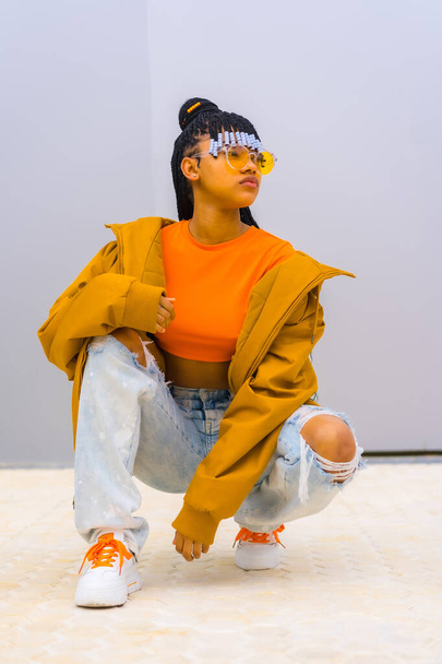 A young trap dancer with braids. Black ethnic girl with t-shirt, orange sunglasses and a jacket, crouched looking at camera with seductive face - Фото, зображення