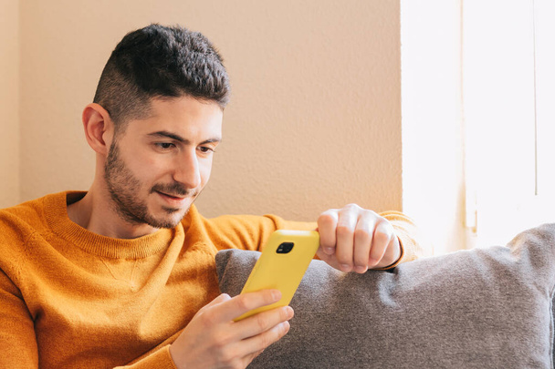 Smiling and happy 30 year old young adult man using his yellow mobile phone on gray sofa indoors - Фото, изображение