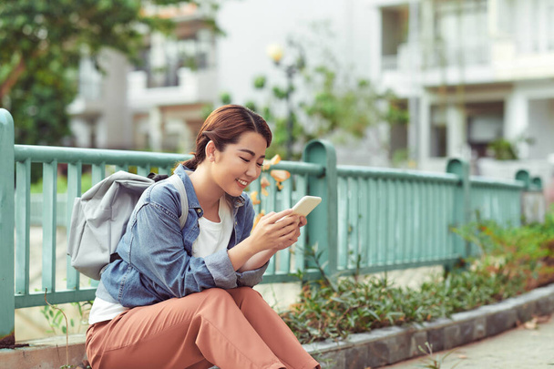 Belle femme dans la ville tenant téléphone mobile, jeune étudiante avec sac à dos regardant la caméra pendant la pause café à l'extérieur - Photo, image