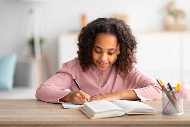 Evde eğitim kavramı. Şirin Afro-Amerikan okul kızı ödev yapıyor ve gülümsüyor, kitapla masada oturuyor. - Fotoğraf, Görsel