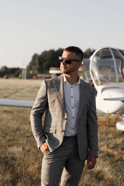 Ein männlicher Pilot vor dem Hintergrund eines Flugzeugs bei Sonnenuntergang. Der Bräutigam im Anzug steht neben seinem Privatflugzeug. - Foto, Bild