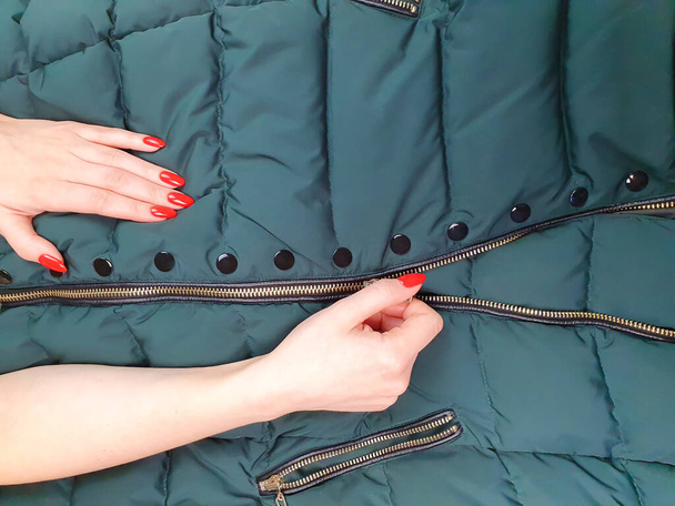 Female hands with red nails close zipper lock on down jacket. Autumn jacket fabric background, green puffer jacket texture. Top view - Photo, Image