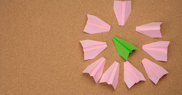 Grünes Papierflugzeug, umgeben von rosafarbenen Papierflugzeugen auf braunem Hintergrund, Draufsicht. Das Konzept der Zusammenarbeit, des Erreichens von Zielen, des Mobbings - Foto, Bild